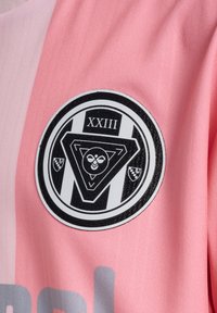 Schwarz-weißes rundes Emblem mit römischer Zahl XXIII und geometrischem Bienendesign auf pink- und hellrosa gestreiftem Stoff.