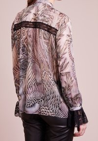 Blusa transparente con un detallado estampado de aves y follaje, con encaje negro en los puños y la parte superior de la espalda, combinada con pantalones de cuero negros.