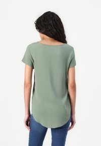 Grüne Kurzarmbluse mit Rundhalsausschnitt, weicher Textur und abgerundetem Saum, kombiniert mit blauen Jeans.