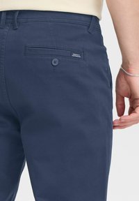 Mornarsko modri bombažni chinos hlače s gladko teksturo, ki imajo zadnji žep z gumbom in majhno oznako z napisom "POSVEČENO NEPOPOLNOSTI."