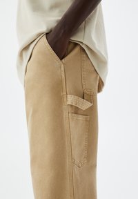 Osoba o ciemnej karnacji ubrana w beżowe spodnie z wieloma kieszeniami i kremową koszulę, ręka swobodnie włożona w przednią kieszeń.