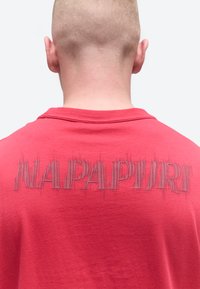 Άτομο με κοντά μαλλιά, που φαίνεται από πίσω, φοράει κόκκινο πουκάμισο με παραμορφωμένο, κατακόρυφο μαύρο κείμενο στην πάνω πλάτη.