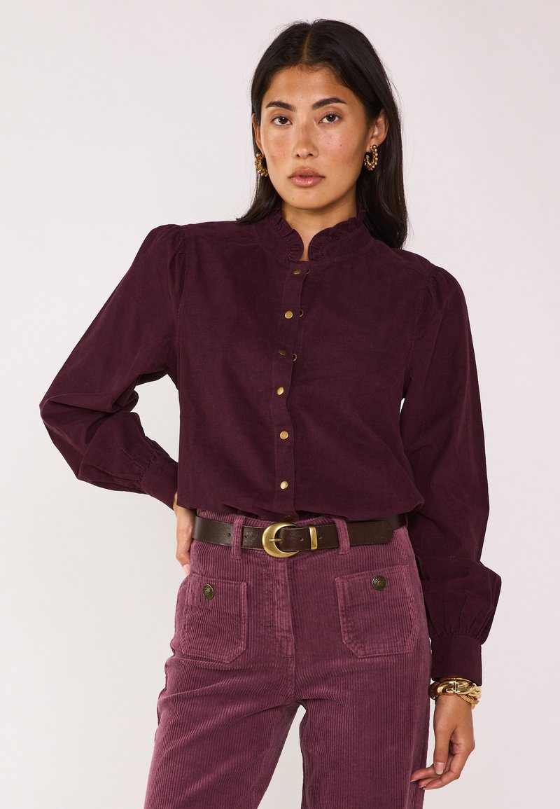 Camicia in corduroy bordeaux con colletto arricciato e bottoni dorati, abbinata a pantaloni coordinati e una cintura marrone scuro con fibbia dorata.