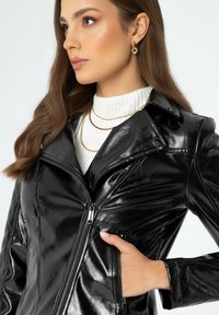 Schwarze Motorradjacke aus Kunstleder mit gesteppten Details, Front-Reißverschluss und seitlichen Taschen. Getragen über einem weißen gerippten Pullover.