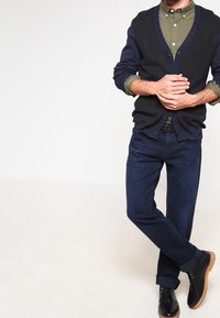 Marinblå cardigan, olivgrön skjorta, mörkblå jeans och svarta stövlar. Cardiganen har en knäppning fram och ribbade mudd. Släta texturer och skräddarsydd passform.