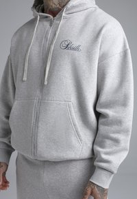 Grijze hoodie met rits, gemaakt van zachte stof, voorzien van een voorzak, trekkoorden en een geborduurd logo op de linkerborst. Textuurafwerking.