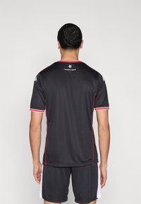 Camiseta deportiva negra de manga corta, con detalles rojos alrededor del cuello y las mangas, y tela texturizada. Presenta un logo en la parte trasera.