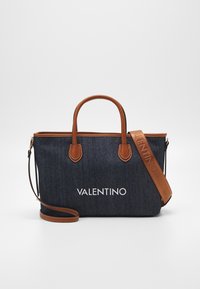 Denim tote väska med tan läderhandtag och axelrem. Har en vit logotyp på framsidan, strukturerad form och kontrasterande sömmar.