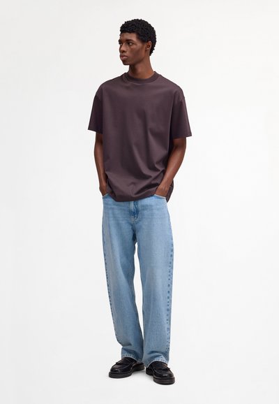 Junger Mann mit dunklem, lockigem Haar, der ein dunkelbraunes, oversized T-Shirt, hellblaue Jeans mit weitem Bein und schwarze Loafers trägt, steht mit den Händen in den Taschen.