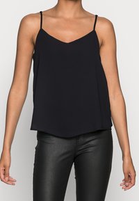 Donna che indossa un top nero senza maniche con scollatura a V e spalline sottili, con pantaloni in pelle nera attillati, in piedi su uno sfondo bianco.