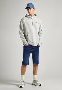 Sudadera gris con capucha y bolsillo frontal, pantalones cortos de mezclilla azul, calcetines blancos y zapatillas grises. La gorra es de color azul claro con texto bordado.