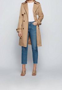 Beżowy płaszcz trencz z dwurzędowym zapięciem, pagonami na ramionach i paskiem w talii, zestawiony z jasnoniebieskimi krótkimi dżinsami i beżowymi szpilkami.