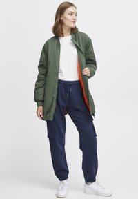 Giacca bomber verde con chiusura a zip e fodera arancione, abbinata a pantaloni della tuta blu navy con tasche laterali. Sneakers bianche completano l'outfit.