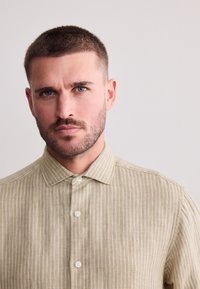 Camicia a righe color cammello con bottoni e colletto. Il tessuto ha una trama testurizzata e presenta bottoni bianchi lungo la parte anteriore.