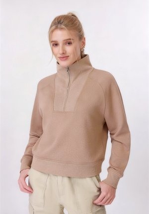 Vrouw met blond haar in een paardenstaart, gekleed in een beige pullover met rits en crème kleurige broek, staand met handen bij de zakken.