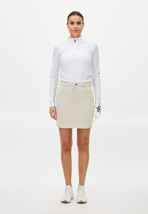 Mujer con una camiseta de manga larga blanca con cremallera, falda beige, guante de golf en la mano izquierda y zapatillas blancas de pie frente a un fondo liso.