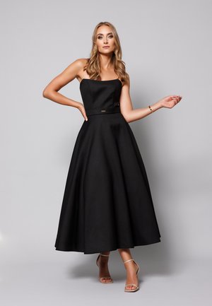 Kobieta z długimi blond włosami, ubrana w czarną sukienkę midi w stylu A bez ramiączek oraz beżowe szpilki na pasku wokół kostki, stoi na tle jednolitego szarego tła.