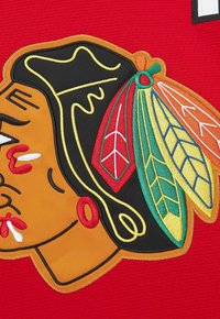 Colorful embroidered Native American head logo with feathers in red, yellow, green, and orange on a red fabric background.