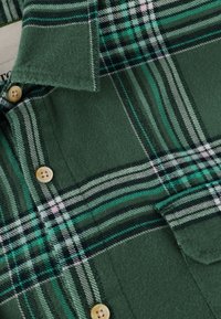 Camisa de franela a cuadros verdes con rayas en tonos de verde oscuro, blanco y negro, acabada con botones marrones y un diseño de bolsillo en el pecho.