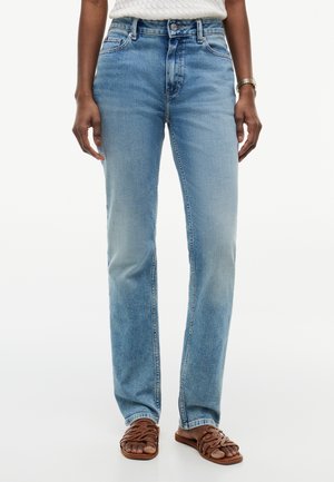 Person iført lyseblå jeans med lige ben, hvid struktureret top, brune sandaler og et guldarmbåndsur, stående mod en hvid baggrund.