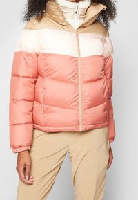 Kurtka puchowa z kolorowym wzorem blokowym w odcieniach brzoskwini, beżu i kremu. Miękki materiał i wysoki kołnierz z zapięciem na zamek.