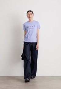 Šviesiai violetinė marškinė su tamsiai mėlynu užrašu "TOMMY JEANS". Dėvimi su aukštu liemeniu tamsiais džinsais. Juoda rankinė ir batai - pasirinktinai.