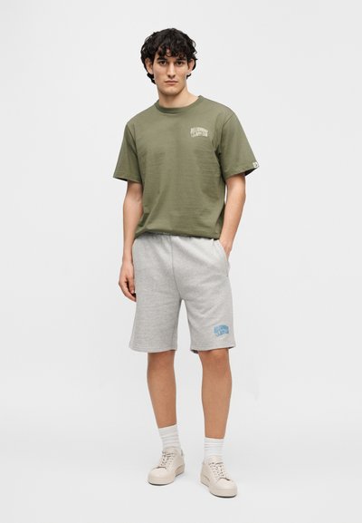 Πράσινο χακί βαμβακερό μπλουζάκι με κοντά μανίκια, σε συνδυασμό με ανοιχτό γκρι βαμβακερά σορτς που έχουν μπλε γραφικό κείμενο. Μπεζ αθλητικά παπούτσια ολοκληρώνουν την εμφάνιση.