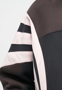 Közelkép egy ujjról, amely fekete és sötétbarna szövetből készült, három világos rózsaszín vízszintes csíkkal és egy függőleges rózsaszín csíkkal a varrás mentén.