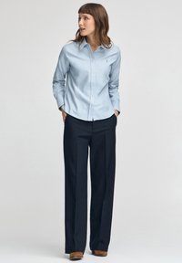 Kvinna som bär en ljusblå skjorta med knappar, svarta vida byxor och bruna skor, stående med händerna i fickorna och tittar åt sidan.