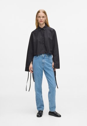 Cămașă neagră scurtă cu mâneci lungi, asortată cu blugi descheiați la talie de culoare albastră deschisă. Pantofii sunt mocasini din piele neagră.