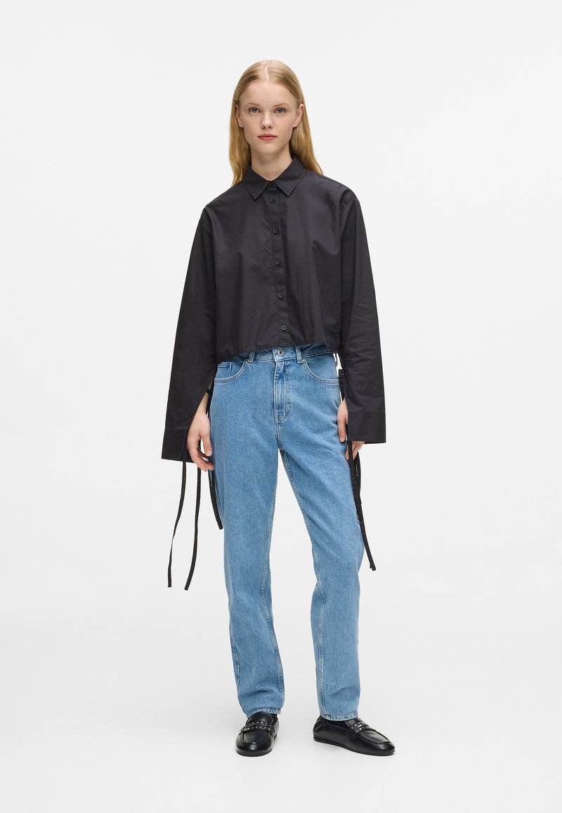 Čierna skrátená košeľa na gombíky s dlhými rukávmi v kombinácii s džínsy svetlomodrej farby s vysokým pásom. Topánky sú čierne kožené mokasíny.