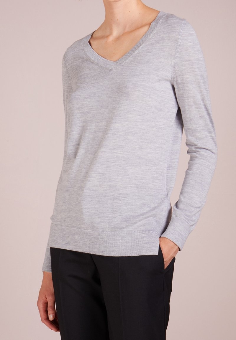 Femme portant un pull léger gris clair à manches longues et col en V, ainsi qu'un pantalon noir, une main dans la poche, sur un fond uni.