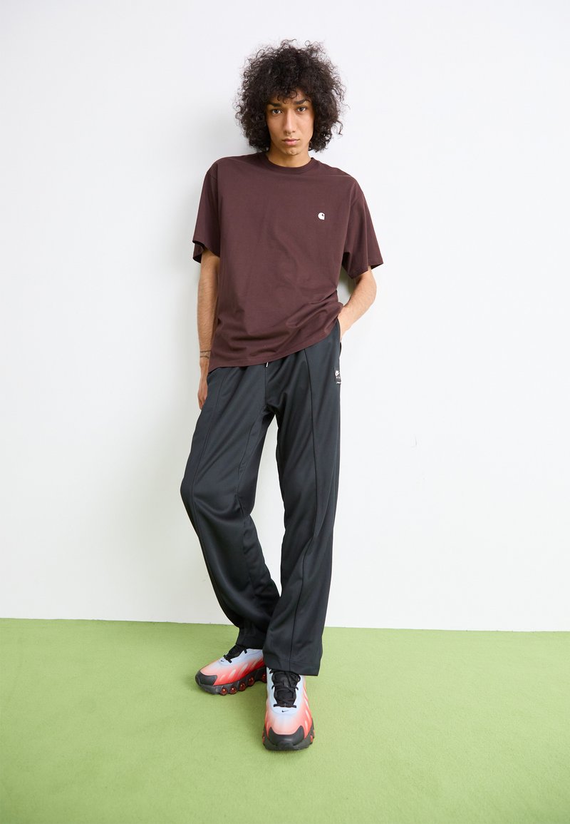 Una persona con capelli ricci sta in piedi contro un muro bianco indossando una maglietta marrone, pantaloni sportivi neri e sneakers bianche-rosse con suola nera.