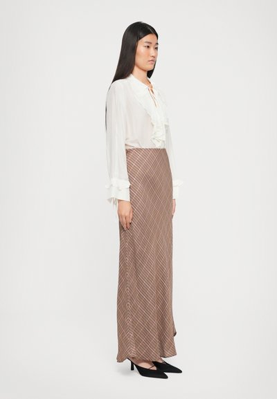 Beige bluse med volanger foran og lange ermer, sammen med et langt brunt skjørt med et diagonalt rutemønster. Svarte hæler.