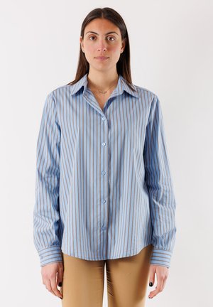 Donna che indossa una camicia a righe verticali blu e marroni con bottoni e pantaloni beige, in piedi davanti a uno sfondo bianco semplice.