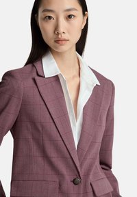 Blazer a quadri bordeaux con rever a punta, chiusura con un bottone e tasche frontali. Indossato sopra una camicia bianca con colletto in tessuto testurizzato.