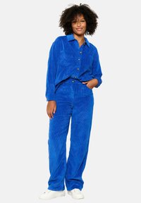 Modra kordurojska kombinacija, ki vključuje srajco na gumb in usklajene hlače. Tkanina ima navpične črte in mehak otip. Bele teniske.