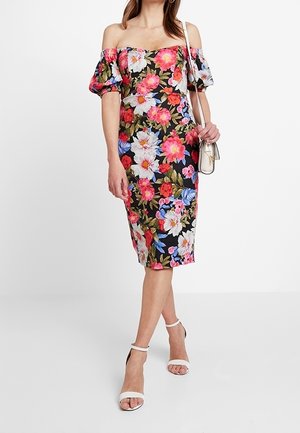 Frau trägt ein schulterfreies Blumenkleid mit pinken, weißen und blauen Blumen, weiße Sandalen mit Knöchelriemen und eine kleine weiße Umhängetasche.