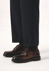 Zapatos de vestir de cuero marrón con un acabado pulido, que presentan cordones y una punta redondeada, combinados con pantalones azul marino y calcetines oscuros.