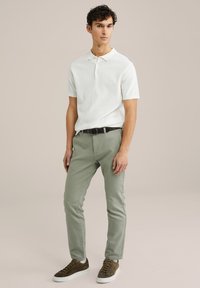 Camisa polo corta de manga blanca texturizada, pantalones ajustados verde claro, y zapatillas de ante marrón con suelas blancas. Un cinturón negro acentúa el conjunto.