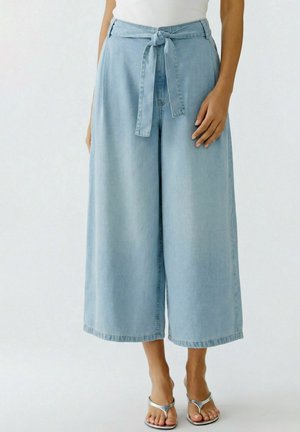 Vrouw die lichtblauwe wijde cropped denimbroek draagt met een gestrikte tailleband en zilveren sandalen met open neus, staand tegen een effen achtergrond.