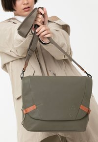 Borsa a tracolla in pelle verde con chiusura a patta, cinturini di accentuazione marroni e tracolla regolabile. Texture liscia e design minimalista.