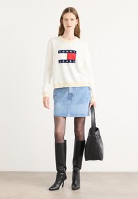 Hvit strikkegenser med "TOMMY JEANS" logo, denim miniskjørt, gjennomsiktige svarte strømpebukser og høye svarte hæler, bærende en svart håndveske.