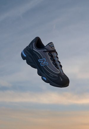 Chaussure de course New Balance gris foncé et bleu suspendue devant un ciel doux du soir en arrière-plan.
