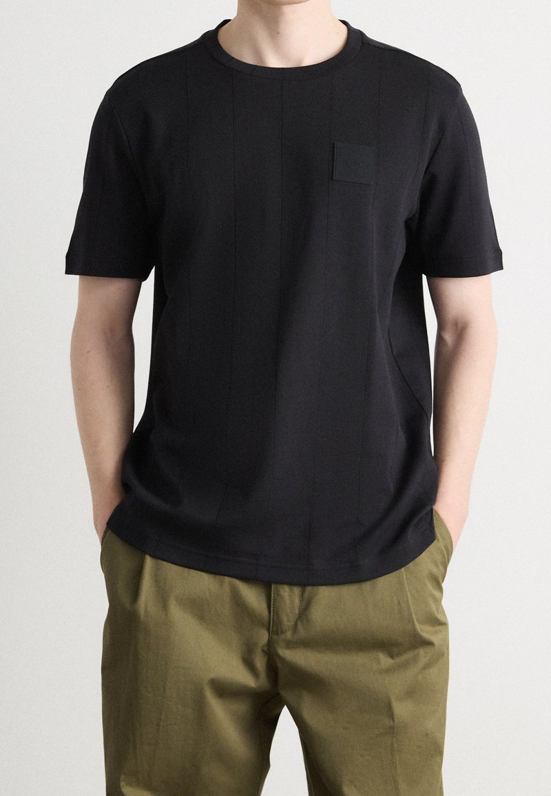 Personne portant un t-shirt noir uni à manches courtes et un pantalon vert olive, les mains dans les poches, debout devant un fond clair.