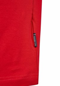 T-shirt rouge en coton à manches courtes, avec une poche latérale et une étiquette de marque bleue avec des lettres blanches près de l'ourlet.