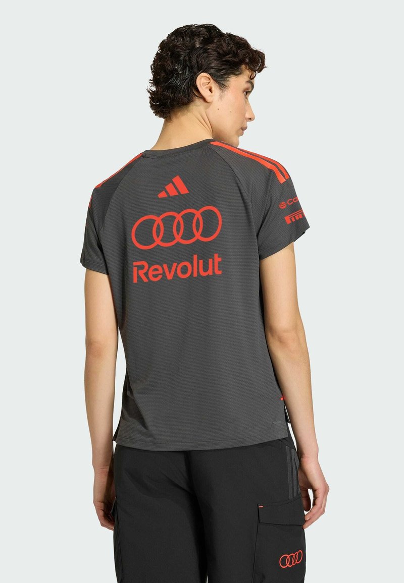 Person med kort krøllet hår iført en grå Adidas sportstrøje med røde logoer, herunder Audi-ringe og Revolut, set bagfra.