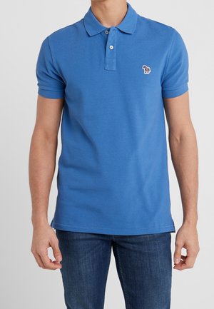 Homme portant un polo à manches courtes bleu avec un petit logo zèbre sur la poitrine gauche, assorti à un jean bleu foncé.
