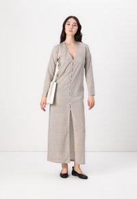 Svetlo šedé dlhé šaty s gombíkmi vpredu, bodkovaným vzorom, dlhými rukávmi a bočným rozparkom. Modelka drží malú bielu kabelku a má na sebe čierne balerínky.