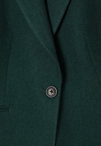 Tessuto verde scuro con motivo a spina di pesce, rever con intaccatura e un unico bottone nero. Presenta profondità testurale e silhouette strutturata.